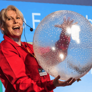 Scientific Sue, wearing a bright red Science2Life lab coat and headset microphone, holds a large transparent balloon filled with tiny white polystyrene beads. The beads cling to the inside surface of the balloon due to static electricity, creating a dramatic "storm in a balloon" effect during a live science show.