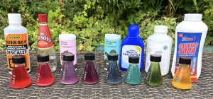 A row of small flasks filled with red cabbage indicator showing a range of colours from red to green, placed in front of household chemicals like vinegar, baking powder, and caustic soda.