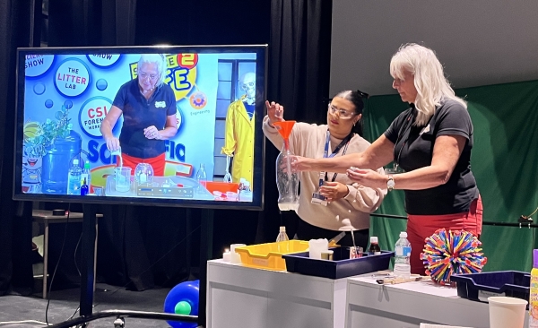 Teacher and pupil demonstrate a science activity at the front of a classroom with a Science of Dragons video playing on a screen behind them.