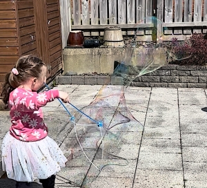 A young girl wearing a pink floral top and white tutu uses a blue tri-string bubble wand to create a large, shimmering bubble outdoors on a sunny day.