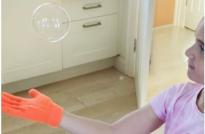 A child wearing an orange glove balances a soap bubble in mid-air inside a bright kitchen.