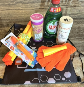 The Bubbleology Science Kit displayed on a marble surface. The kit includes Fairy Liquid, bicarbonate of soda, xanthan gum, a bubble blower, orange gloves, yellow bubble wands, a tri-string rope wand, and a black Science2Life drawstring bag with a chemistry-themed design.