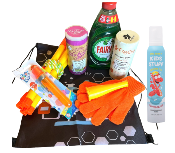 The Bubbleology Science Kit displayed on a surface. The kit includes Fairy Liquid, bicarbonate of soda, xanthan gum, a bubble blower, orange gloves, yellow bubble wands, a tri-string rope wand, Bouncing foam soap and a black Science2Life drawstring bag with a chemistry-themed design.