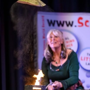 Scientific Sue performs a fiery grain-dust explosion demonstration on stage, with a burst of flame rising from a small box as she speaks to the audience.