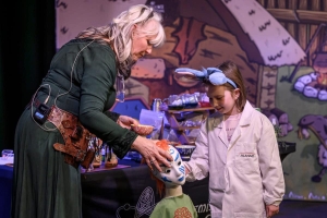 Scientific Sue and a young volunteer in a lab coat place a bandage on a mannequin styled as a Celtic warrior during a live science show.