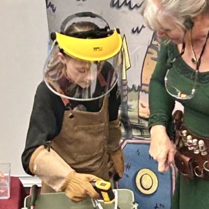 A young volunteer dressed as a blacksmith, wearing protective gloves and a face shield, uses a tool while Scientific Sue guides them during a Celtic coin casting demonstration.