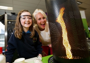 A child wearing safety goggles smiles beside an adult as they watch a tall, swirling flame rising inside a perforated metal cylinder during a science demonstration.