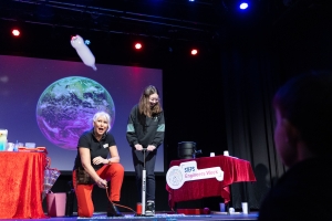 A presenter kneels beside a student using a hand pump on stage, launching a plastic bottle that shoots upward through the air, illustrating the force created by high pressure. A projected image of Earth is visible behind them.